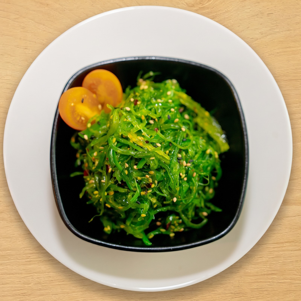 Bright green seaweed salad with sesame seeds and two halved cherry tomatoes in a black bowl.