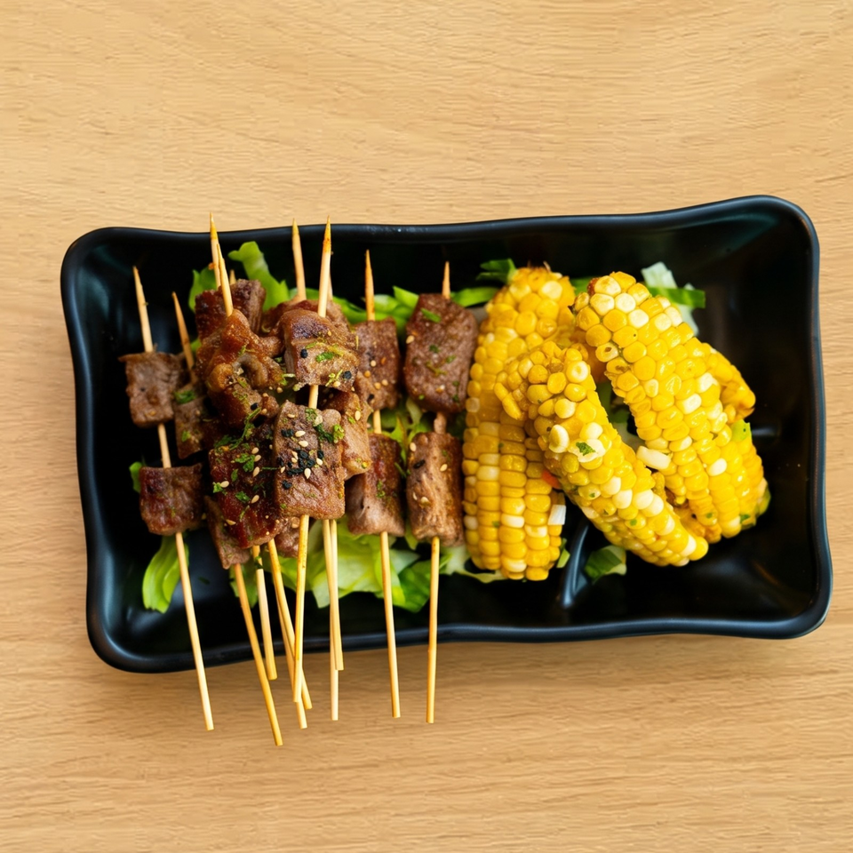 Beef skewers with sesame seeds and herbs, alongside corn on the cob, on a black plate.